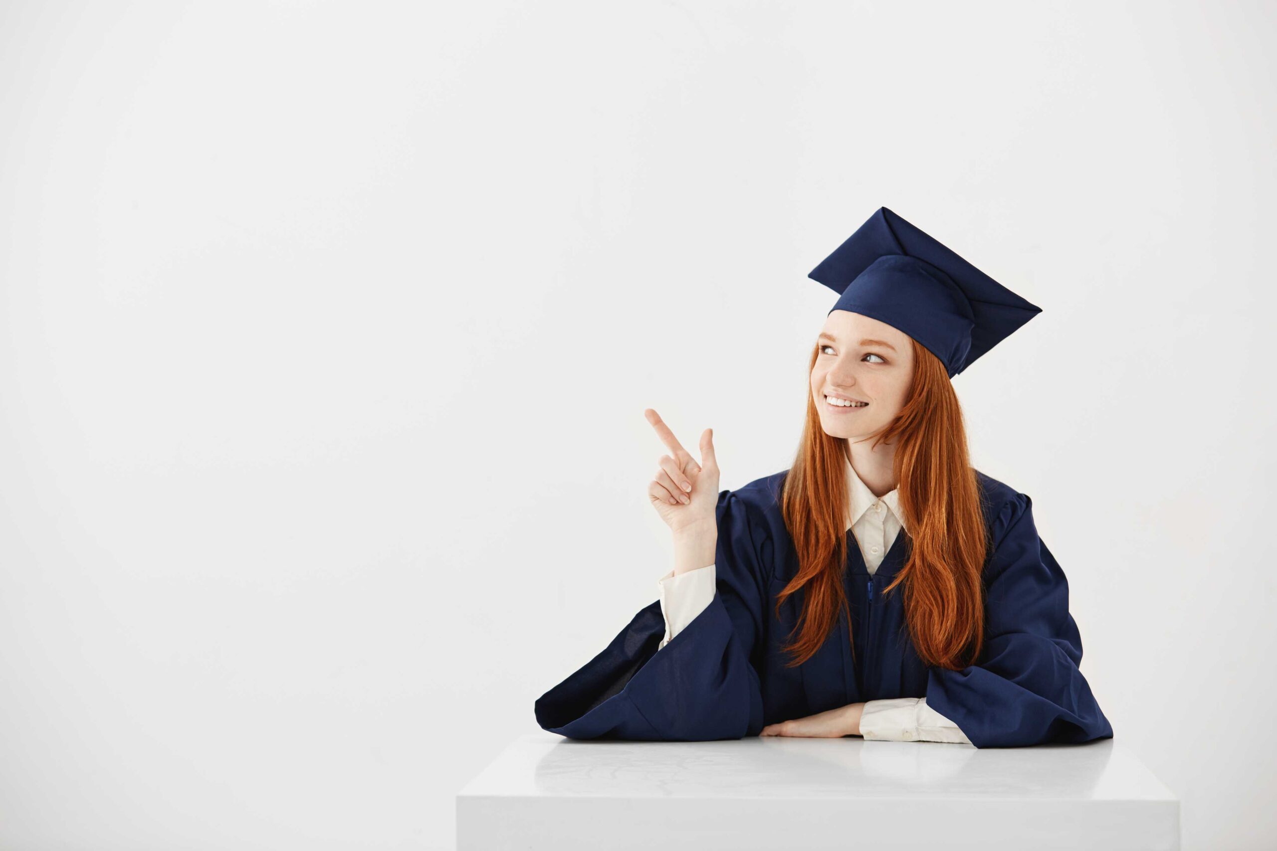 young-female-university-graduate-academic-cap-sitting-table-smiling-pointing-left-future-lawyer-engineer-showing-idea (1)