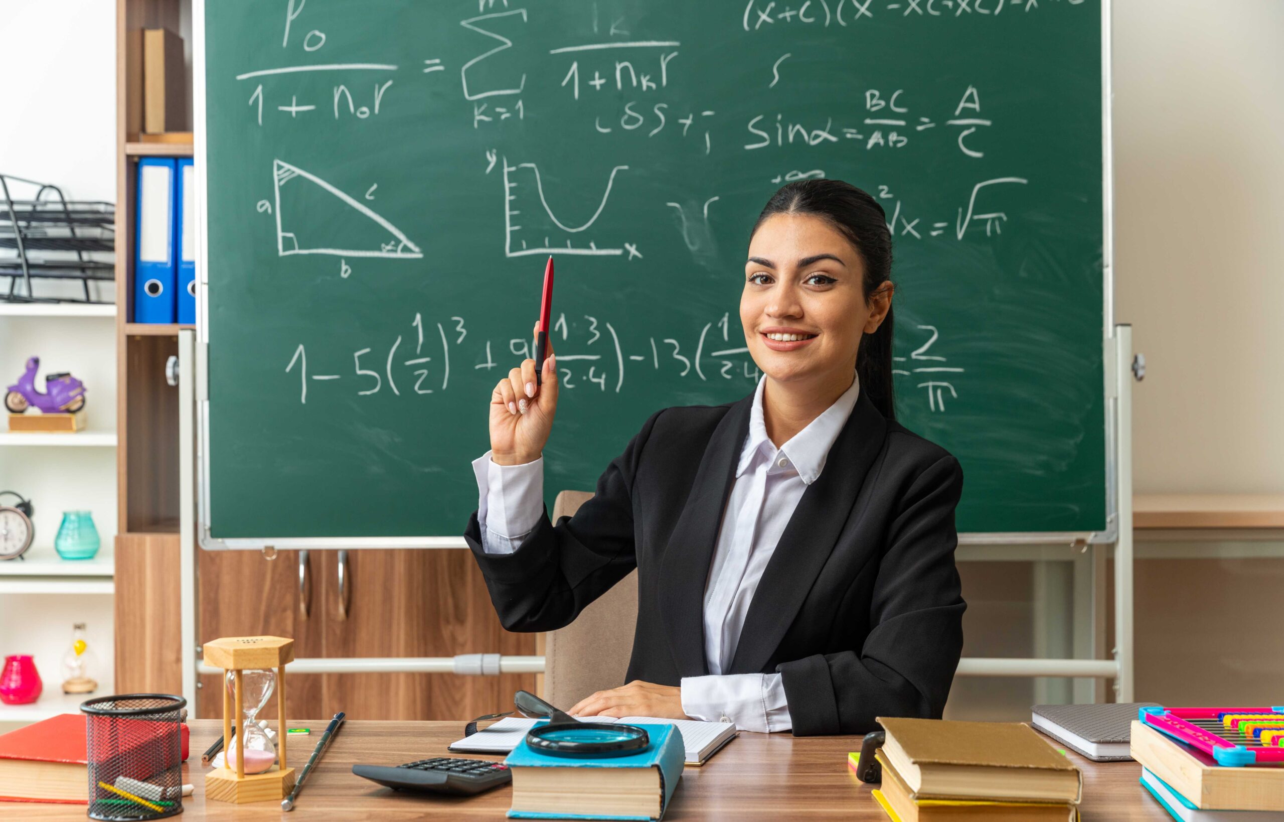 impressed-young-female-teacher-sits-table-with-school-tools-raising-pen-classroom